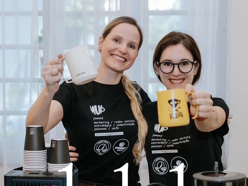 Gabi e Kay brindando com canecas atrás da estação de café